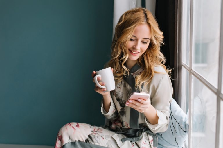 fille assise sur le rebord de la fenêtre avec une tasse de café, du thé et un smartphone dans les mains