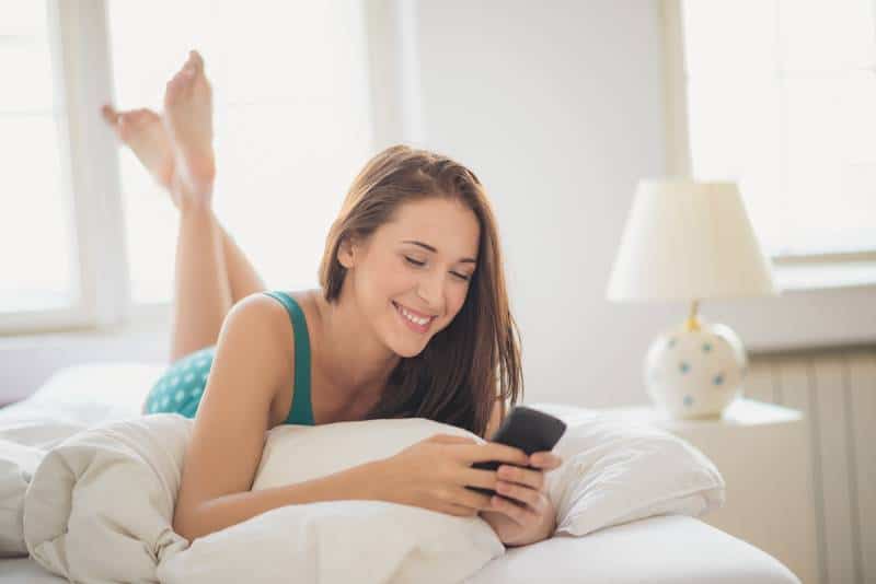 une brune souriante tapant sur son téléphone alors qu'elle est allongée sur son lit