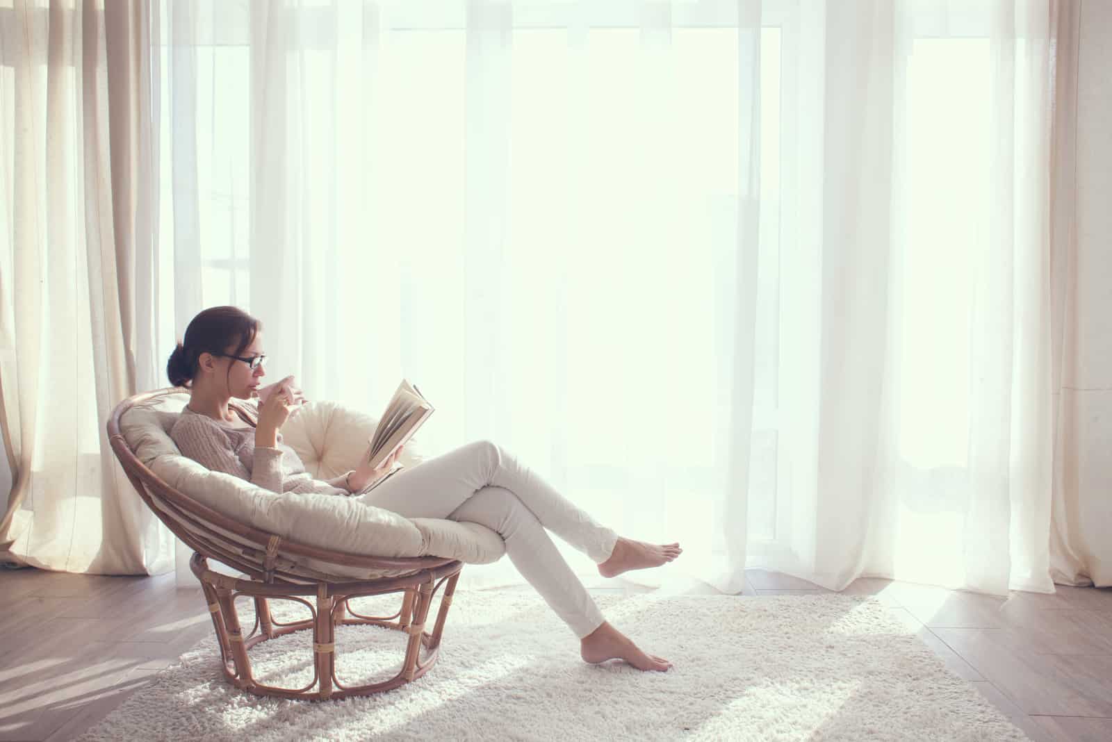 la femme est assise dans un fauteuil en lisant un livre et en buvant du café