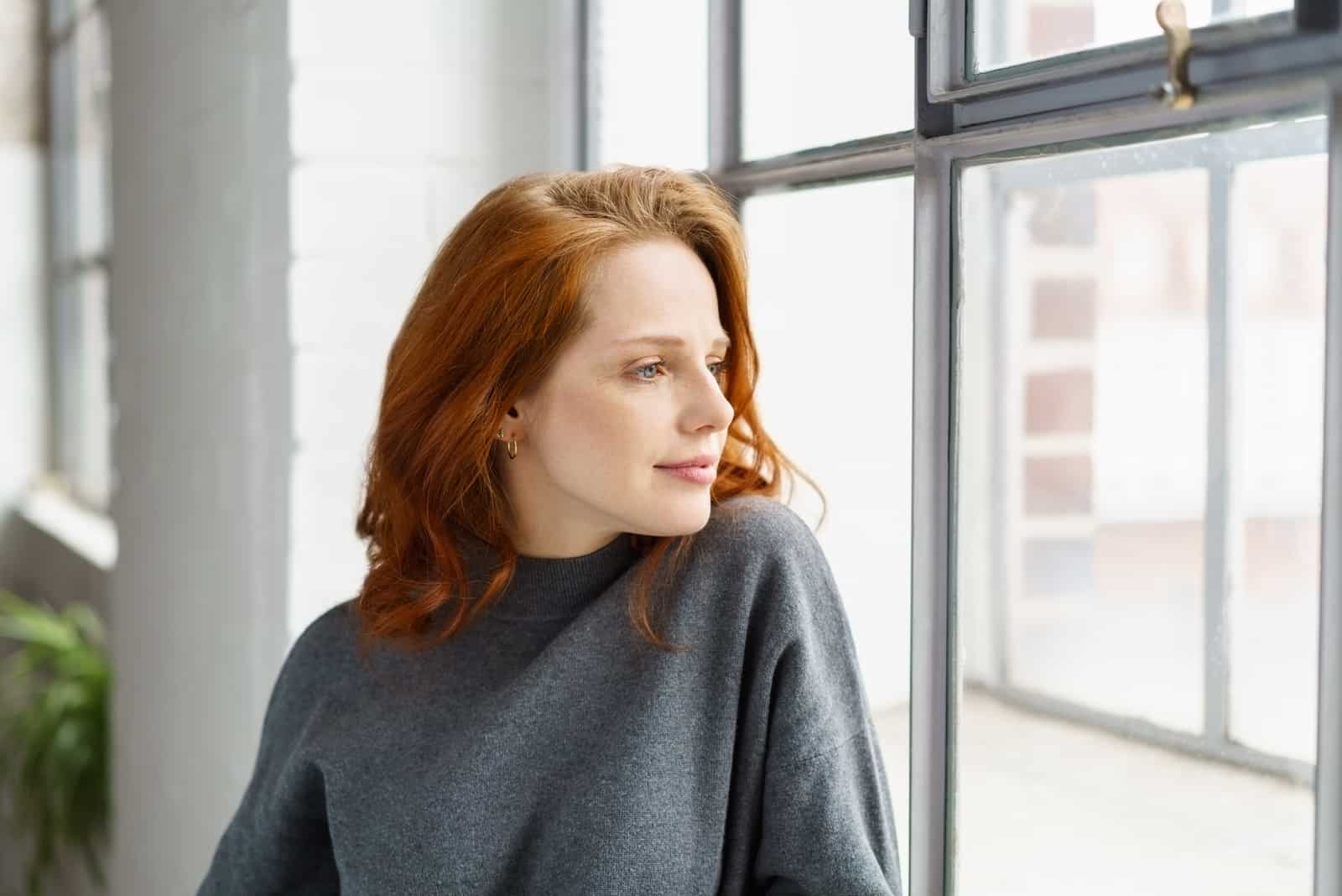 Pensive séduisante jeune femme rousse debout appuyé sur le rebord à regarder par la fenêtre