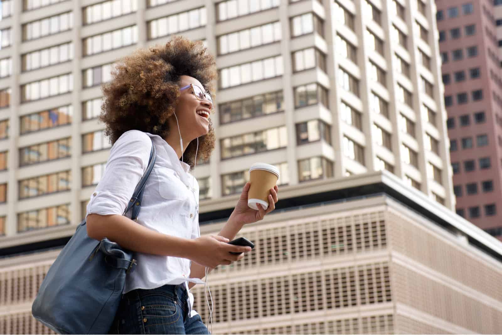 Une femme marche dans la rue avec du café à la main et des écouteurs dans ses oreilles