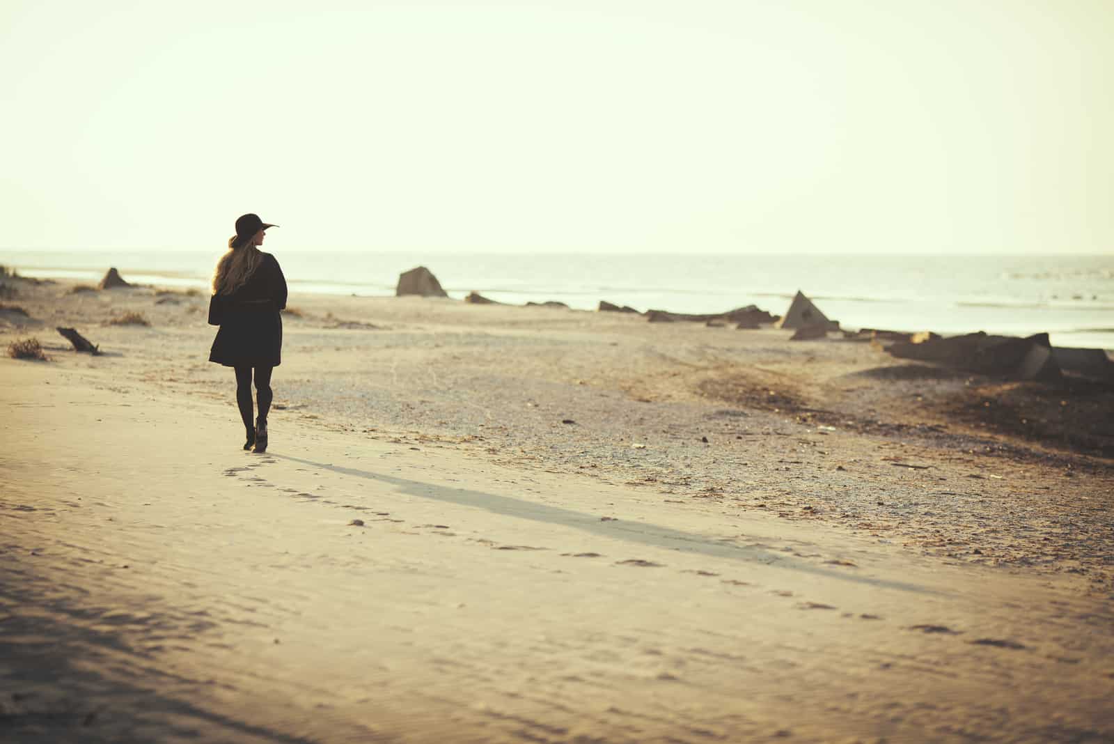 la femme marche sur la plage avec un chapeau et un manteau noir