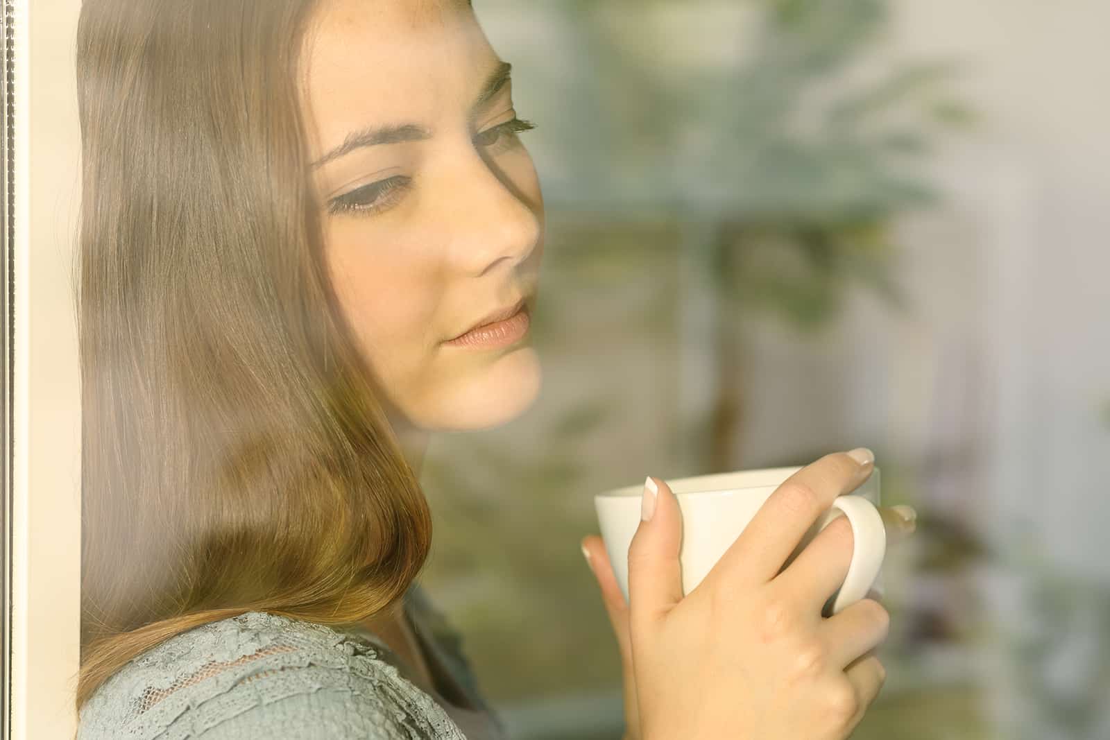 Dame pensive regardant à travers une fenêtre et tenant une tasse de café