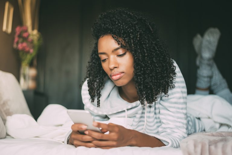 Une femme sérieuse aux cheveux bouclés à l'aide de smartphone sur le lit