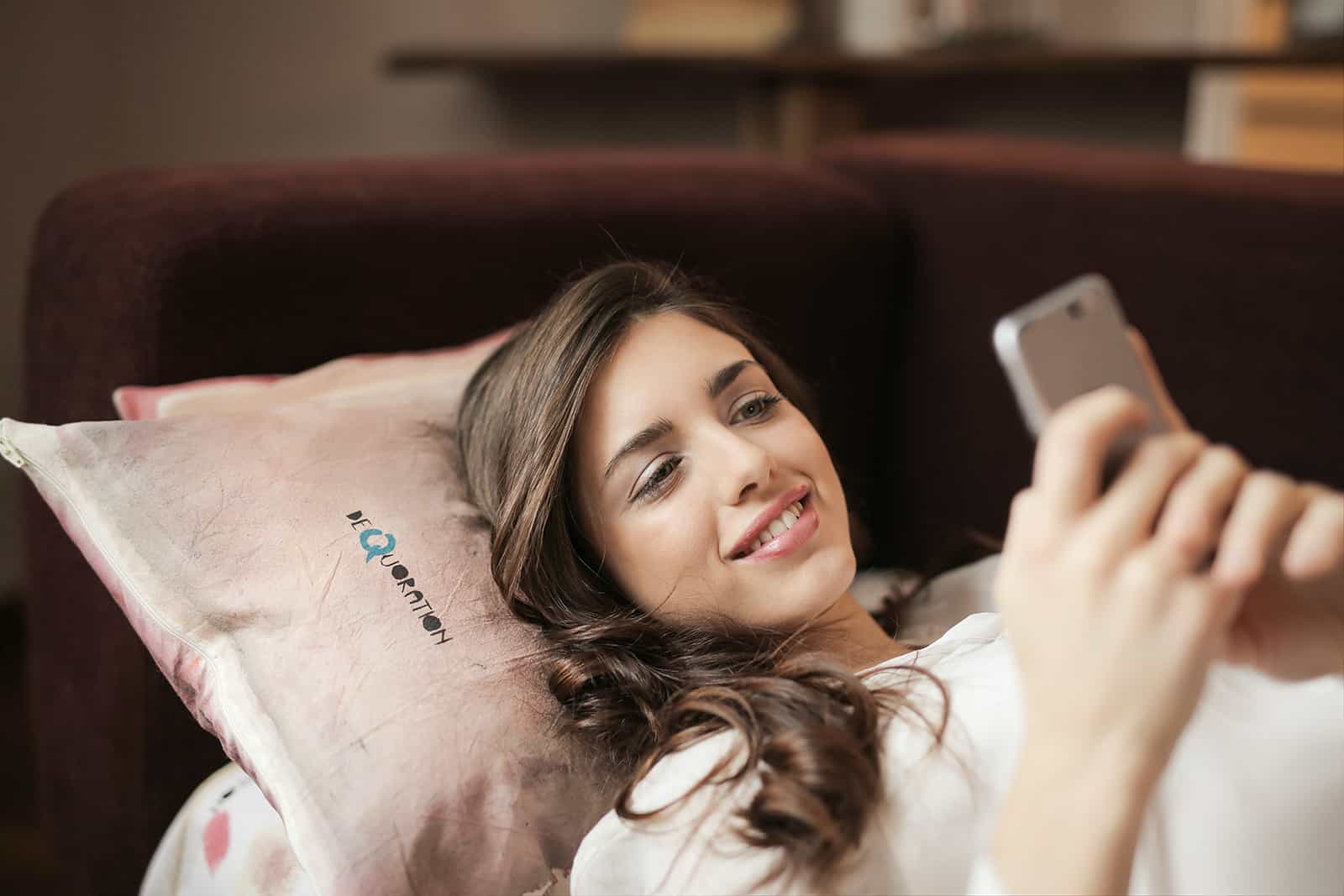 une femme souriante regardant smartphone en position couchée sur le canapé