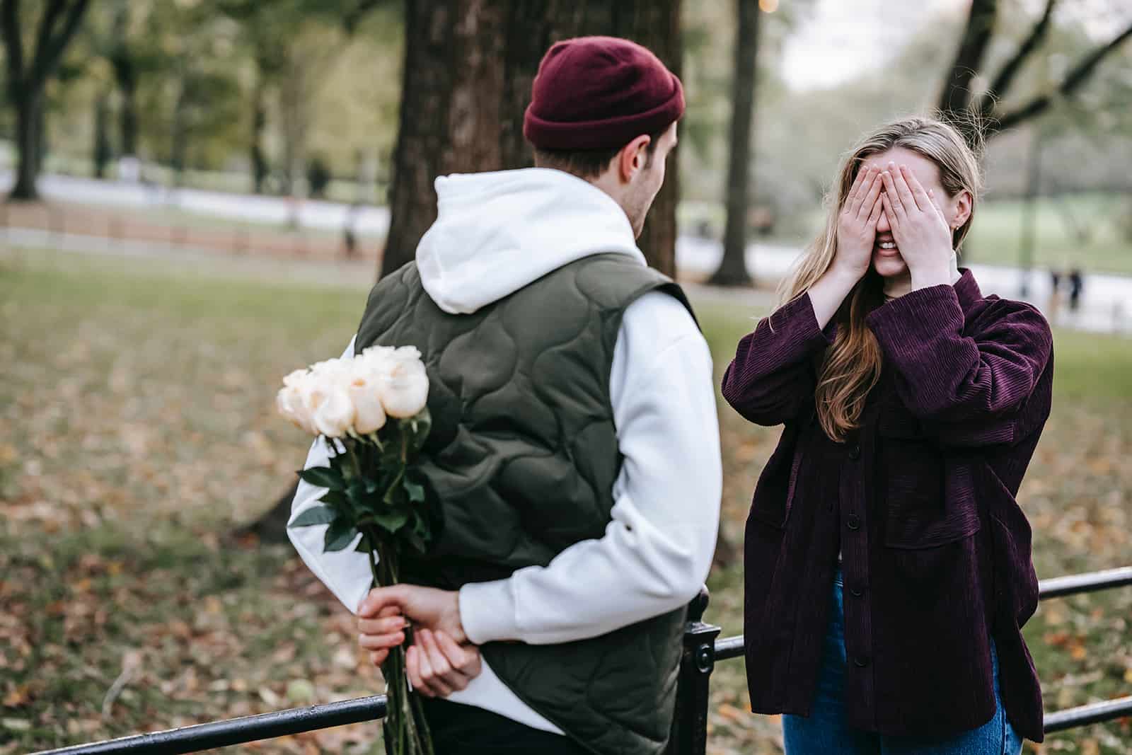 Un homme tenant un bouquet de roses derrière son dos devant une femme couvrant ses yeux avec des paumes