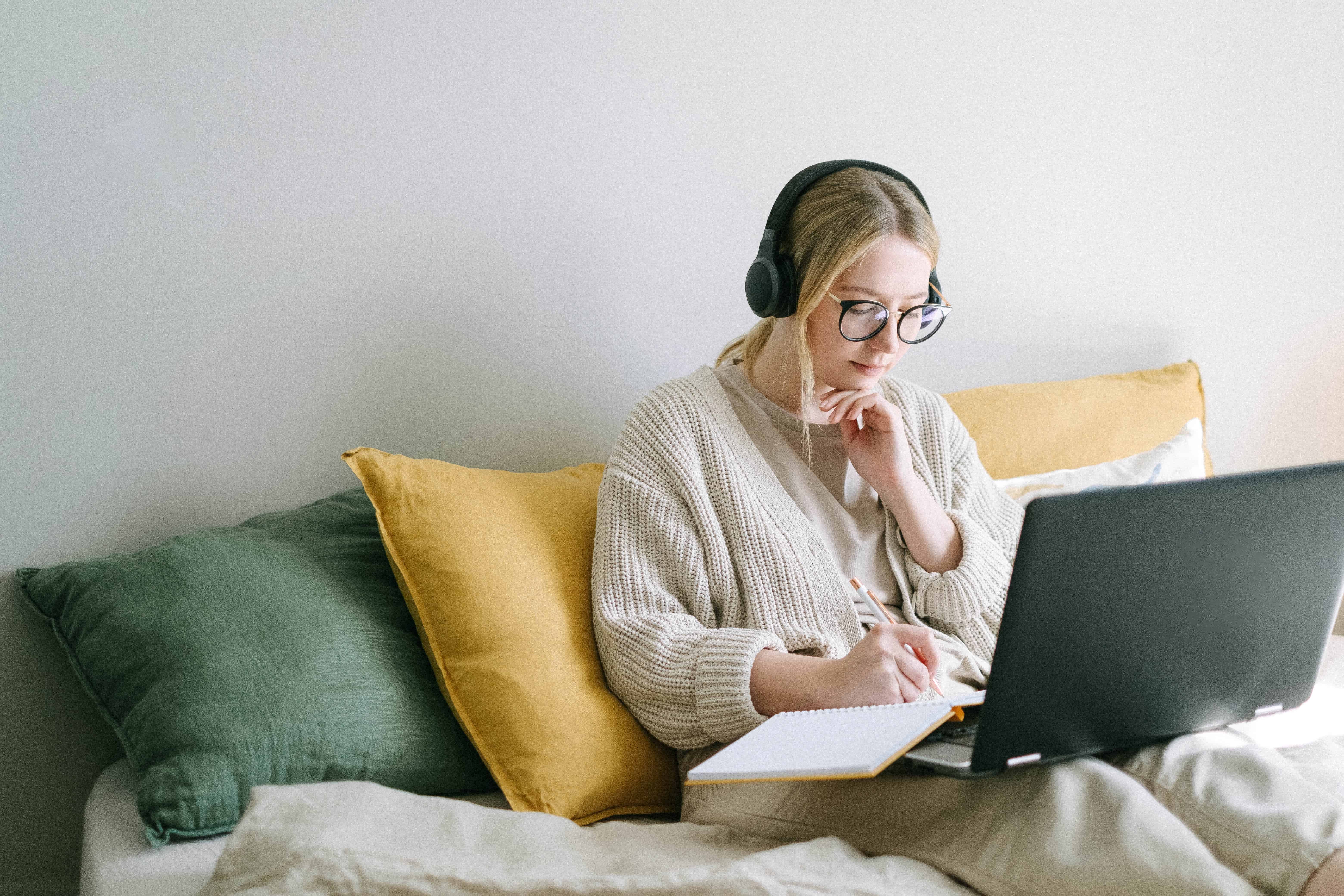 Une femme avec des écouteurs en prenant des notes tout en utilisant un ordinateur portable dans le lit