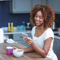 une femme souriante tient le téléphone tout en prenant le petit déjeuner