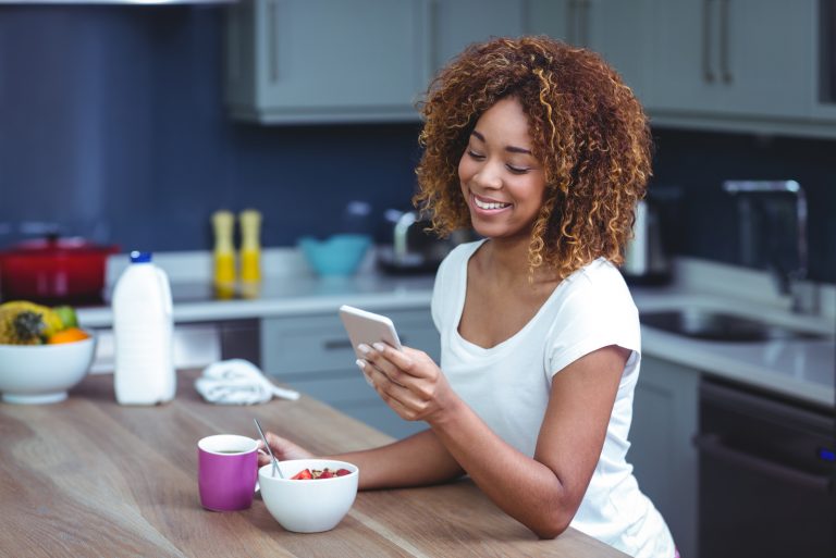 une femme souriante tient le téléphone tout en prenant le petit déjeuner