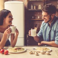 Beau jeune couple parle, se regarde et sourit en mangeant et en buvant dans la cuisine à la maison