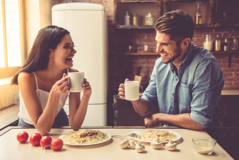 Beau jeune couple parle, se regarde et sourit en mangeant et en buvant dans la cuisine à la maison