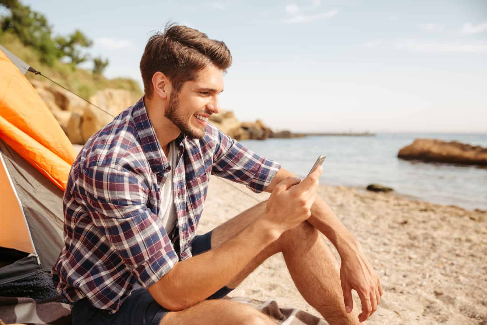 Souriant jeune homme touriste à l'aide de smartphone assis dans une tente touristique à la plage