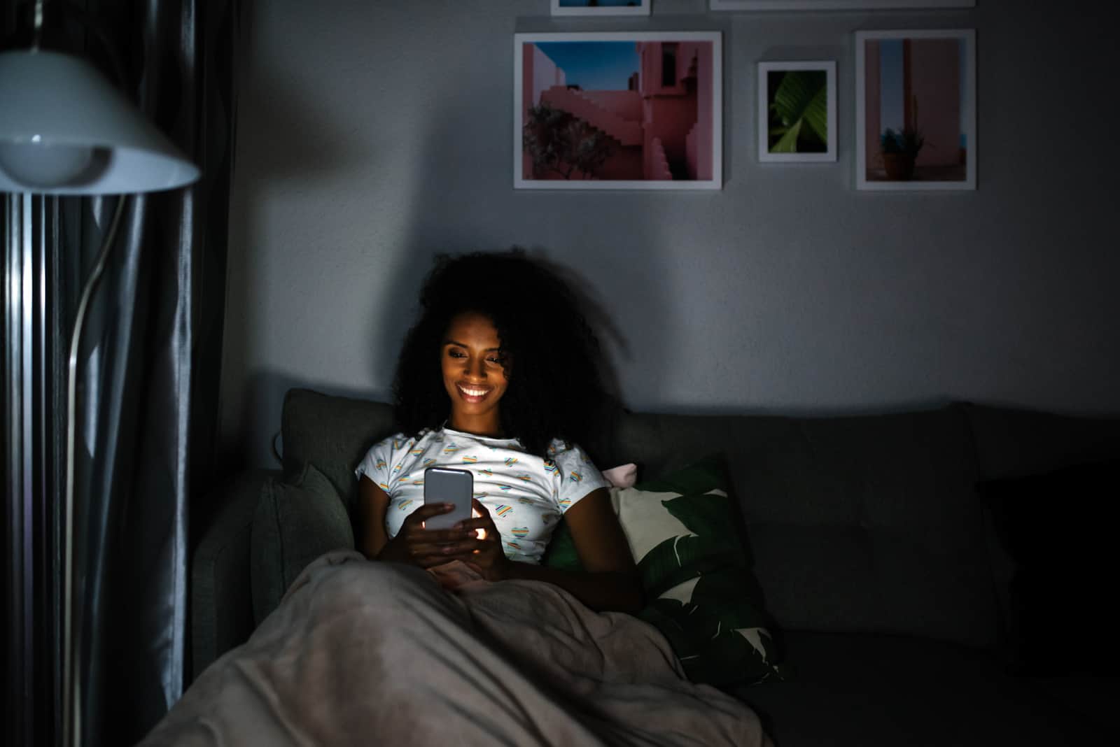 Une femme souriante aux cheveux crépus se trouve dans son lit et tient un téléphone dans sa main