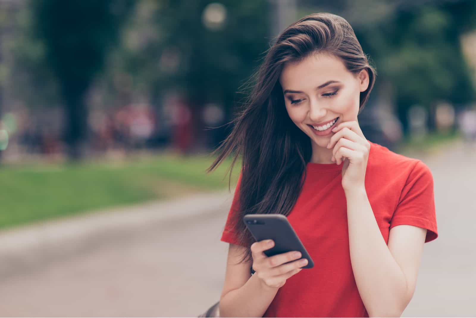 Une femme souriante aux longs cheveux bruns se dresse dans la rue tenant un téléphone à la main