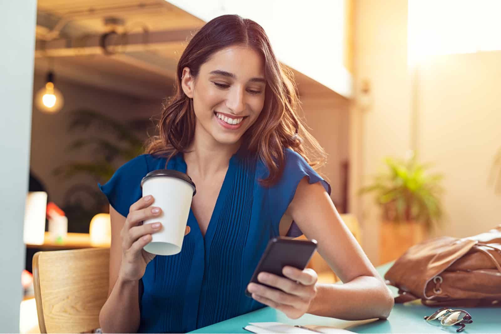 Une jeune femme souriante buvant du café tenant un téléphone à la main et en riant