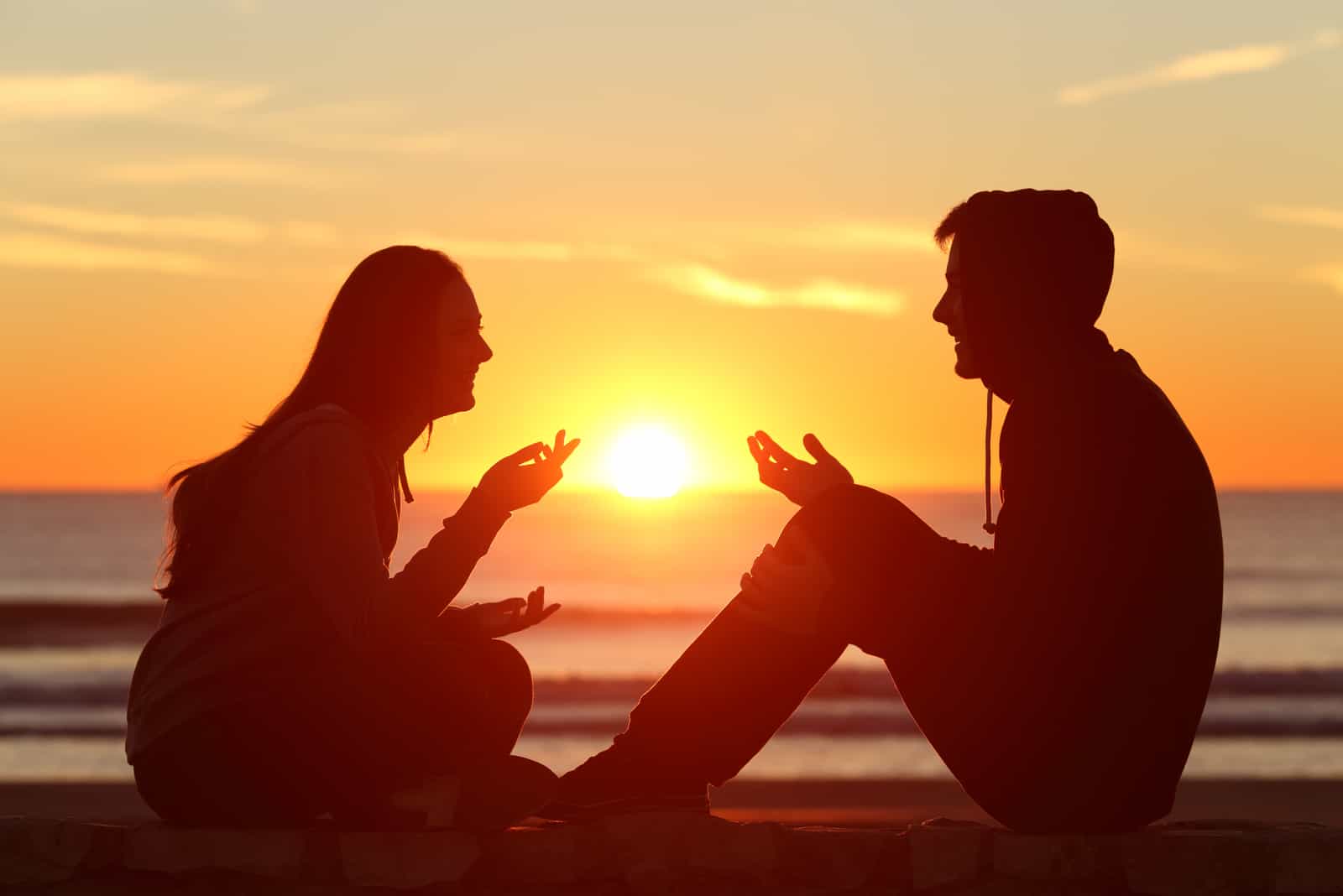 Vue latérale d'un couple d'adolescents assis et parler au lever du soleil sur la plage avec le soleil au milieu