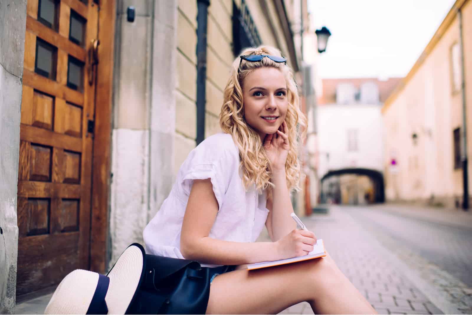 une femme aux cheveux blonds est assise sur le trottoir et écrit