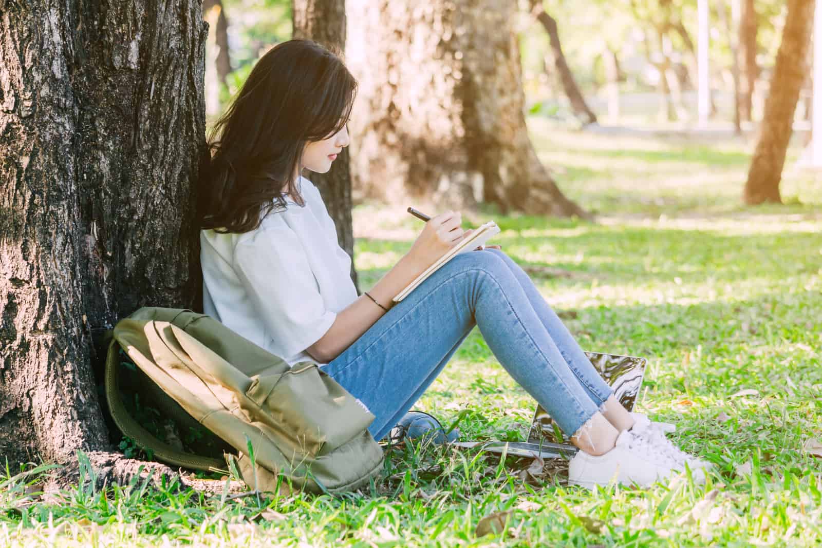 une femme est assise sur l'herbe appuyée contre un arbre et écrit une lettre