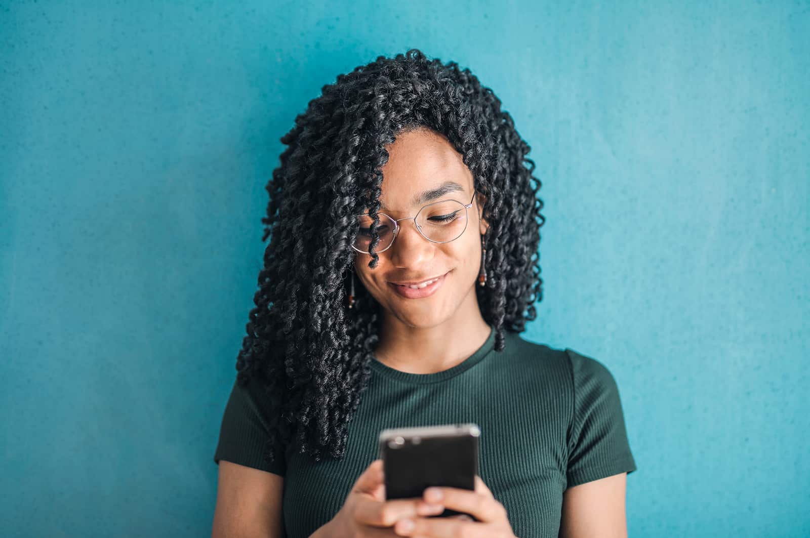 Une femme souriante aux cheveux bouclés en regardant un smartphone et debout près du mur bleu