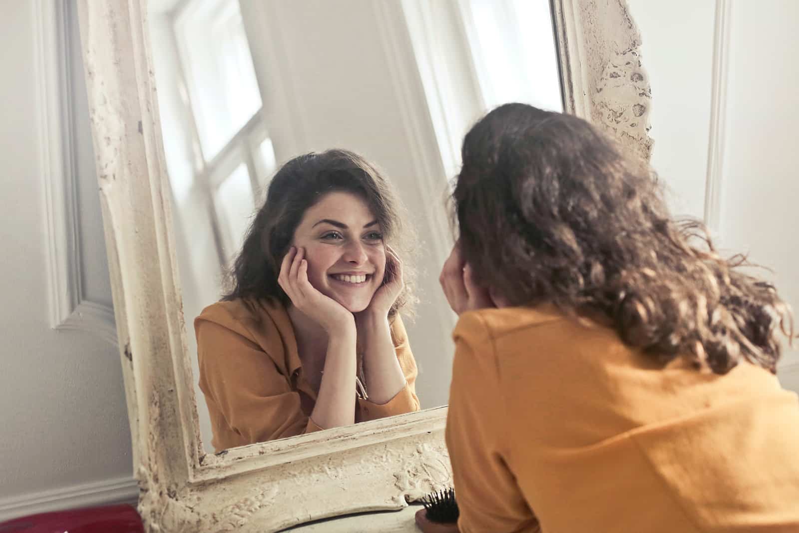 Une femme souriante se regardant dans le miroir tout en appuyant une tête sur ses paumes
