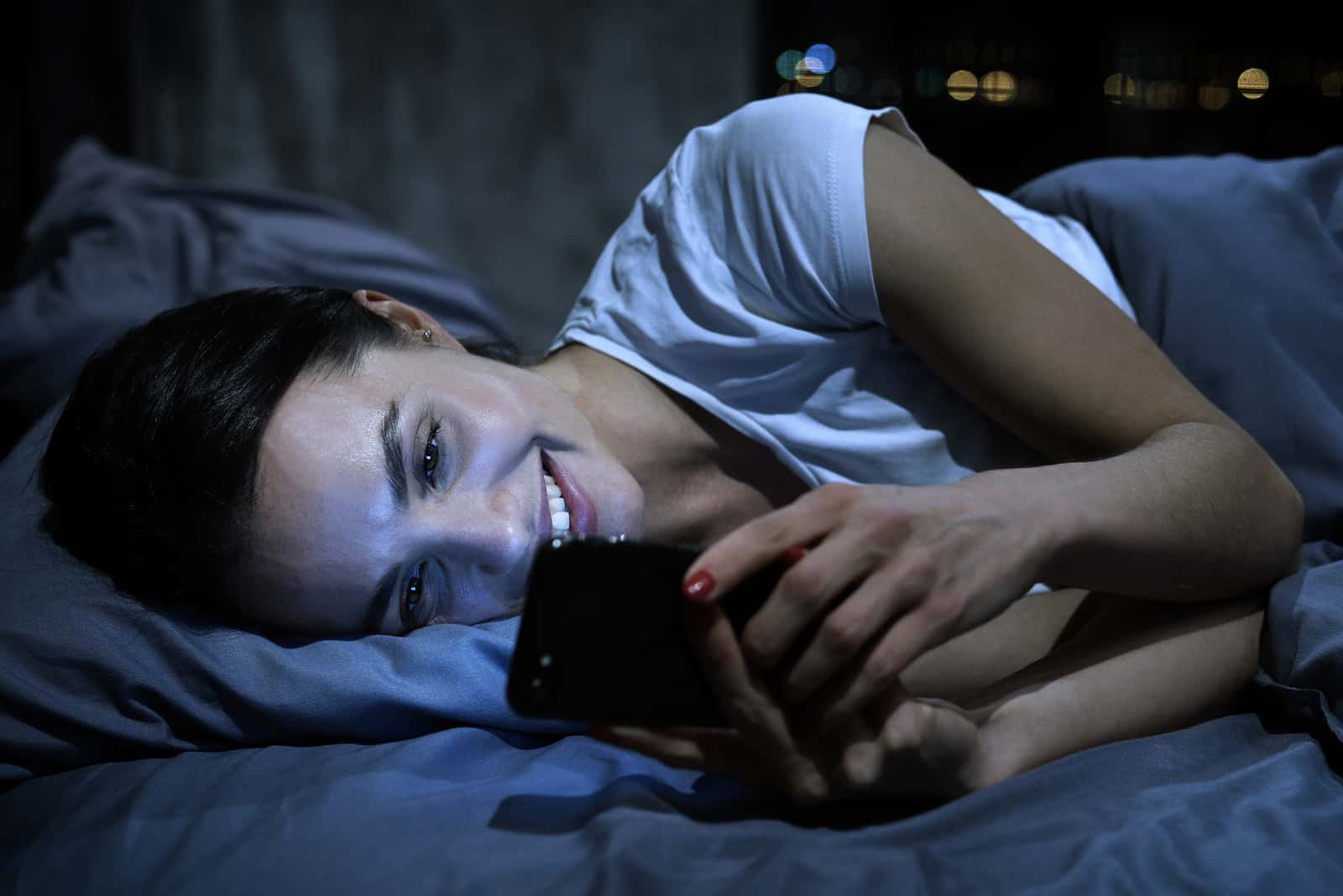 une femme souriante se trouve dans son lit et tient un téléphone dans sa main
