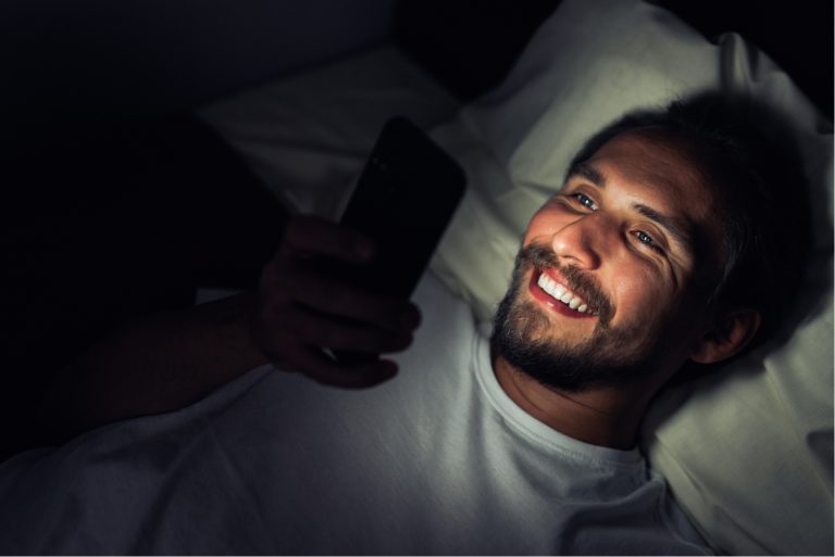 un homme souriant couché dans son lit et en appuyant sur un téléphone