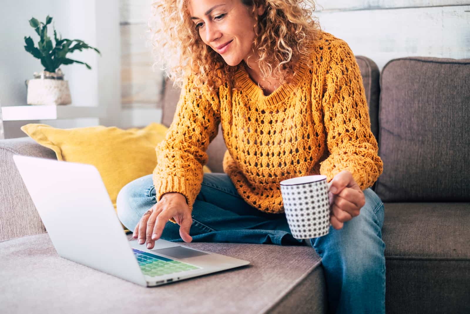Une femme est assise sur un bouton de canapé sur un ordinateur portable et boit du café