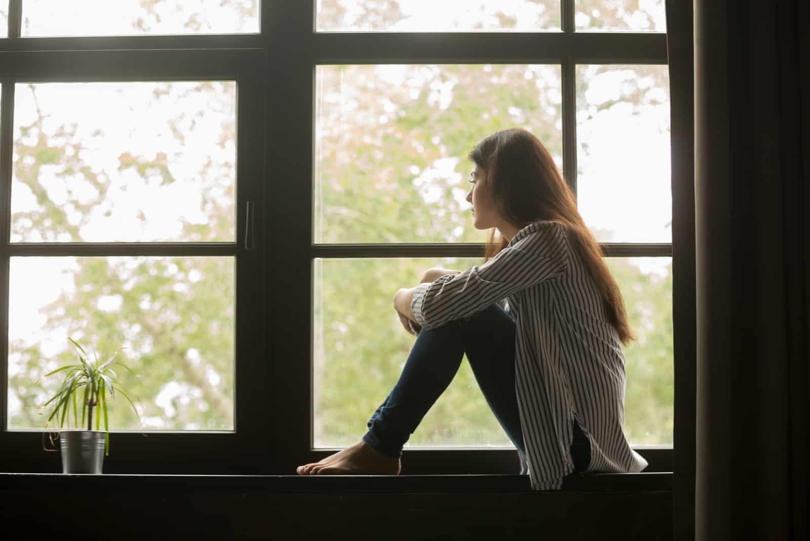 fille assise sur le rebord embrassant les genoux regardant la fenêtre