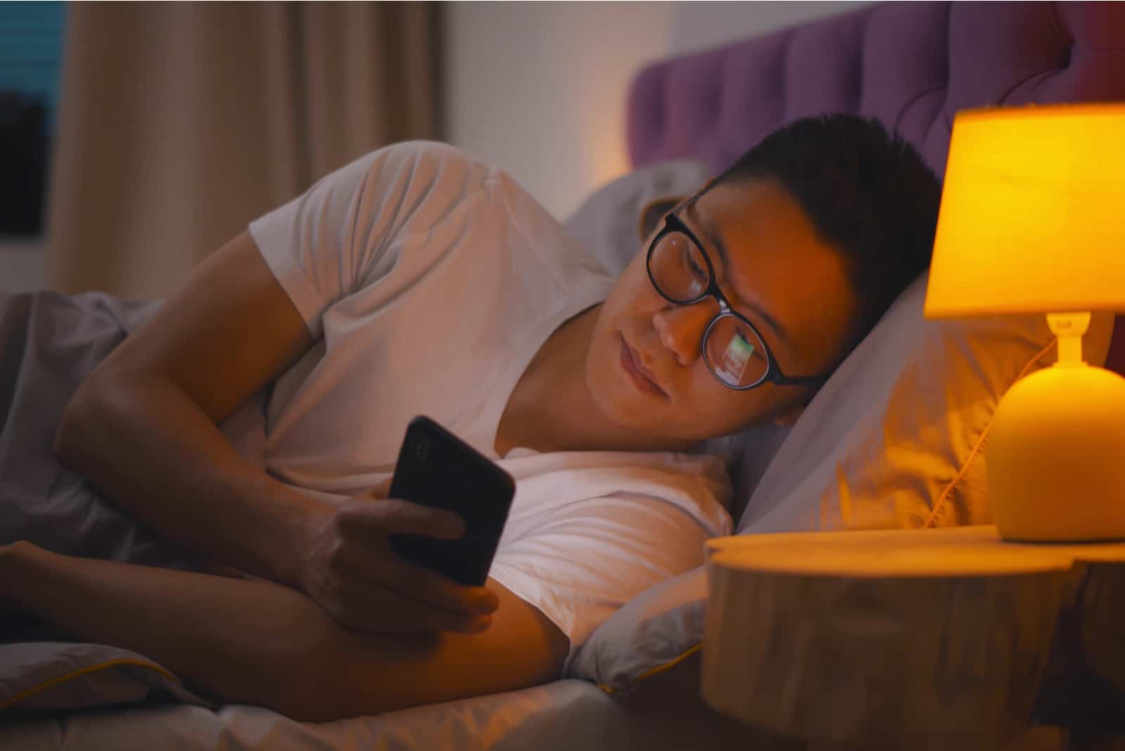 un homme avec des lunettes se trouve dans son lit et tient un téléphone dans sa main