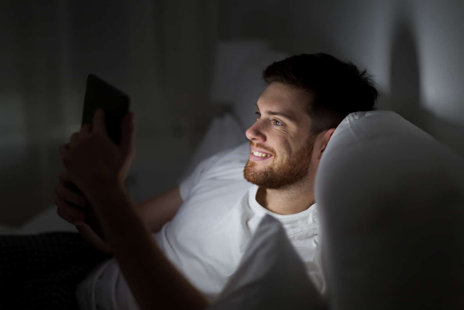 un homme souriant couché dans son lit et en appuyant sur un téléphone