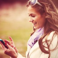 Femme souriante lisant un message sur un téléphone intelligent, prise de vue en extérieur, couleurs rétro.