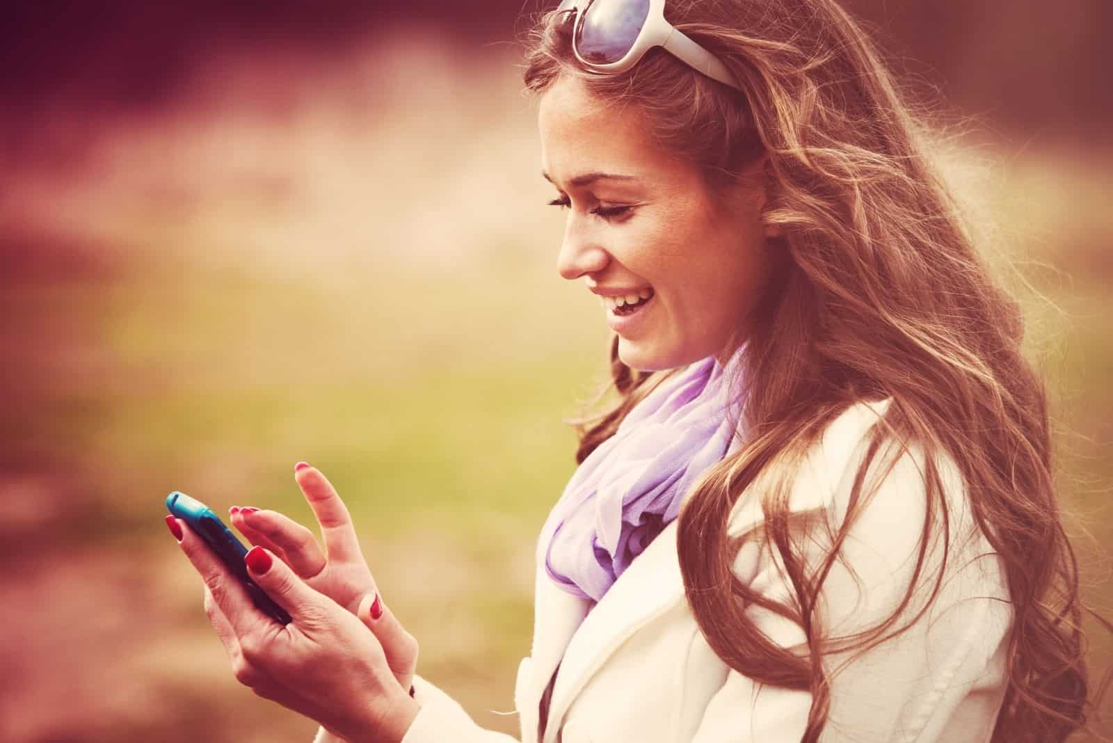 Femme souriante lisant un message sur un téléphone intelligent, prise de vue en extérieur, couleurs rétro.