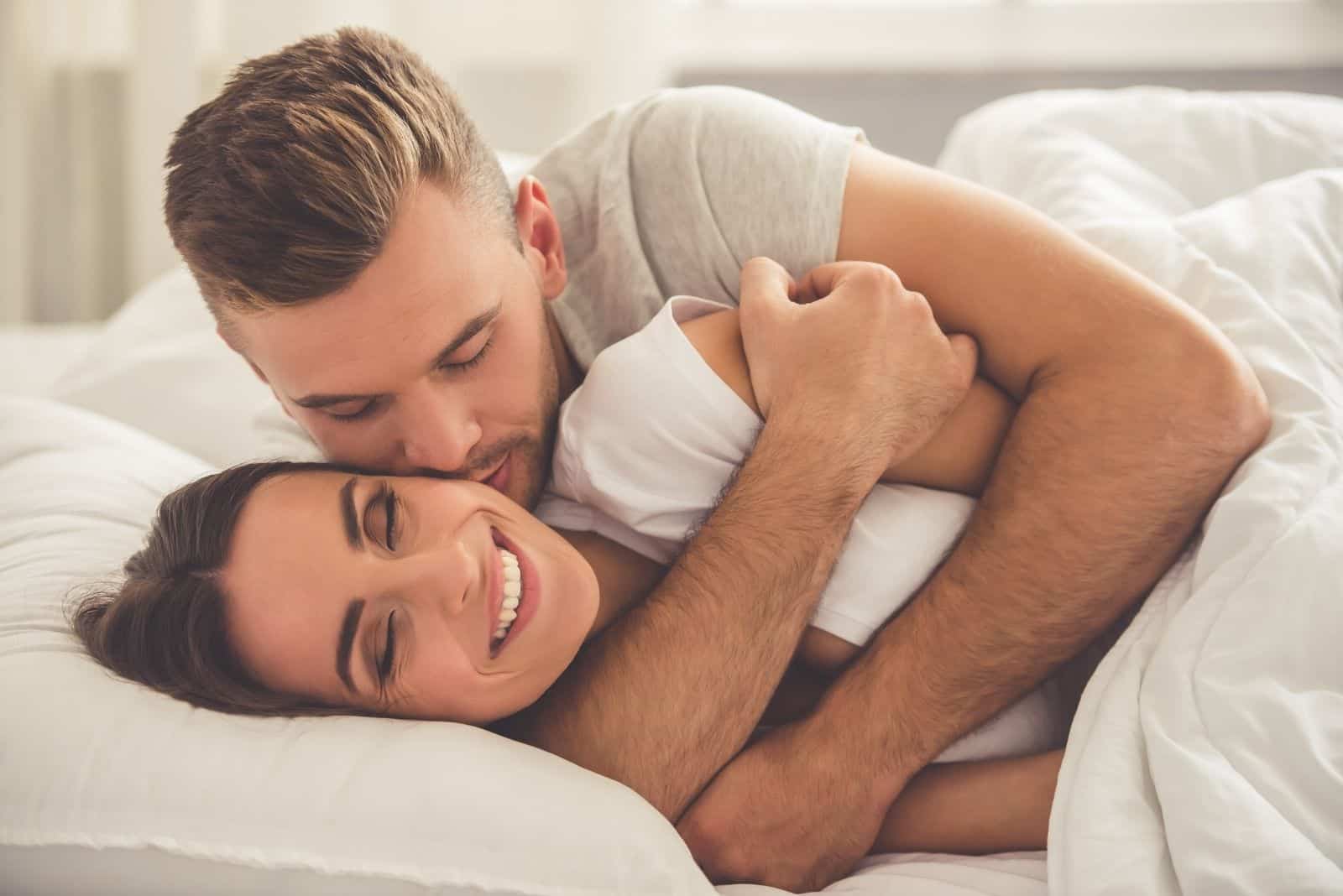 Un beau jeune homme serre dans ses bras et embrasse sa belle épouse souriante sur la joue alors qu'ils sont tous deux allongés dans le lit le matin.