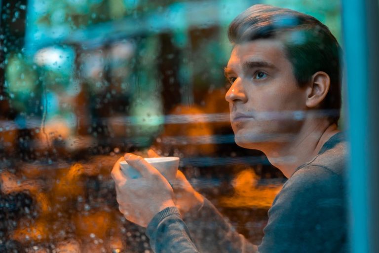 Bel homme buvant du thé ou du café un jour de pluie et regardant par la fenêtre