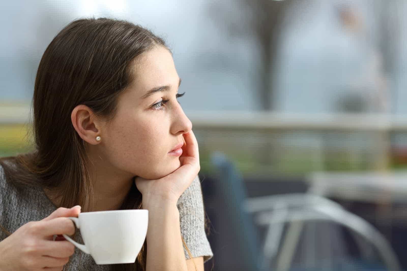 Femme pensive tenant une tasse de café en détournant les yeux dans une terrasse de restaurant en un jour de pluie