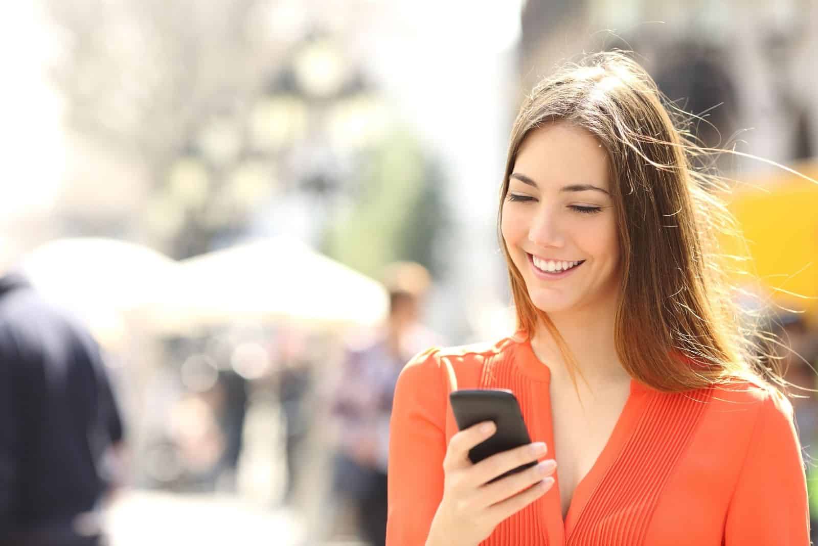 Femme portant une chemise orange en train d'envoyer un texto sur un téléphone intelligent en marchant dans la rue par une journée ensoleillée.