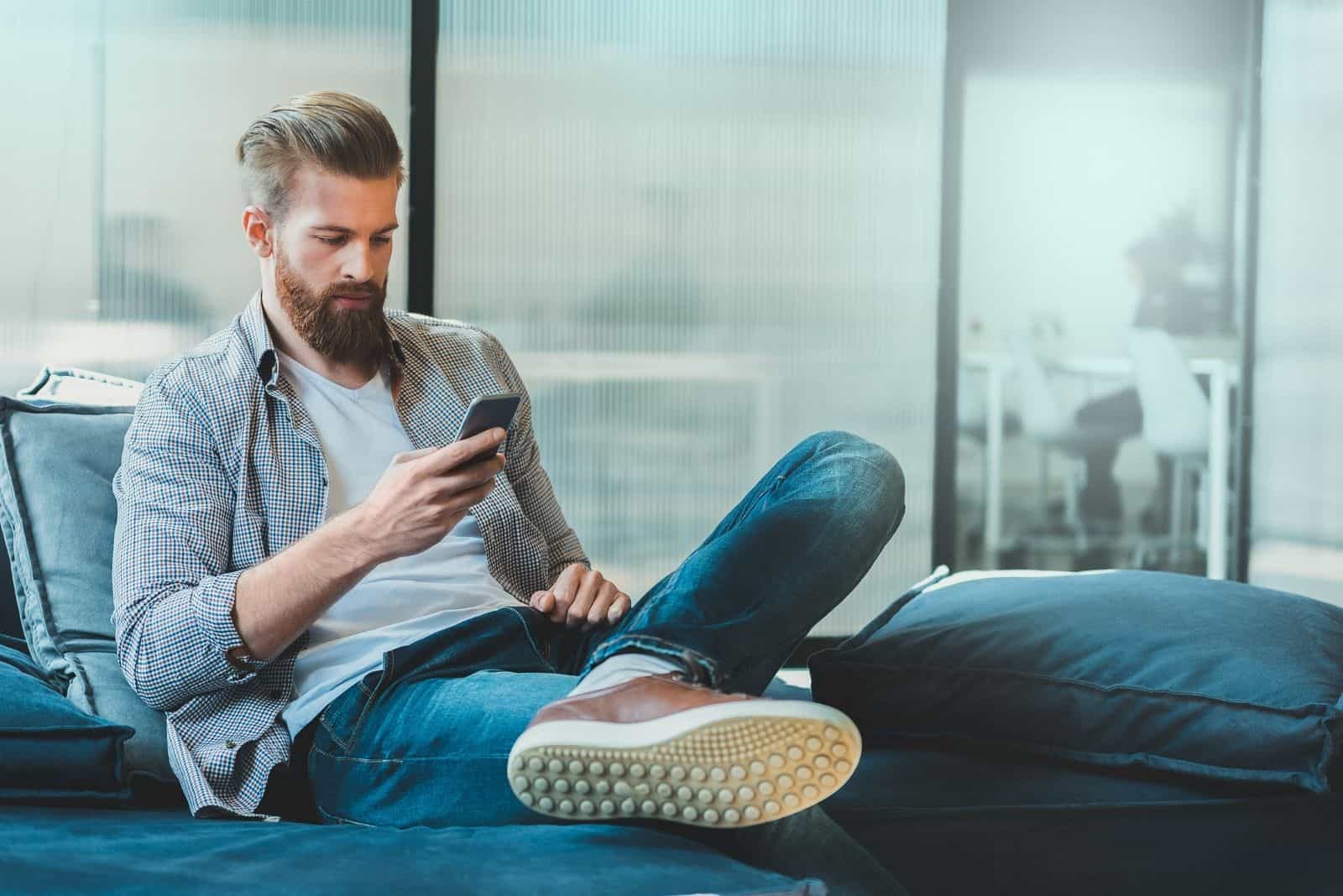 Un homme barbu confiant est assis sur un canapé et tape sur smartphone.