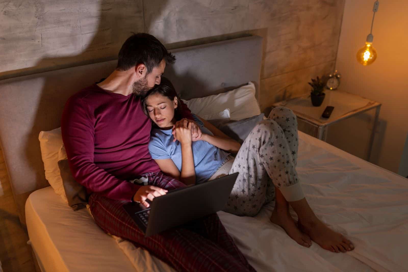 Homme travaillant tard dans la nuit, assis dans son lit et utilisant un ordinateur portable pendant que sa femme dort à côté de lui.
