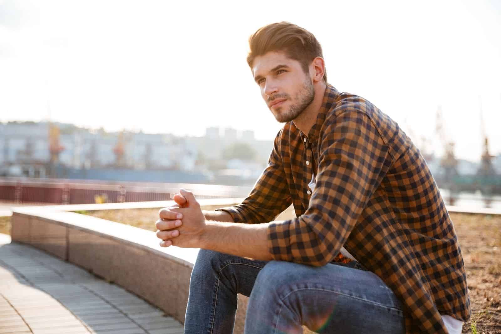 Pensive jeune homme en chemise à carreaux assis et penser à l'extérieur