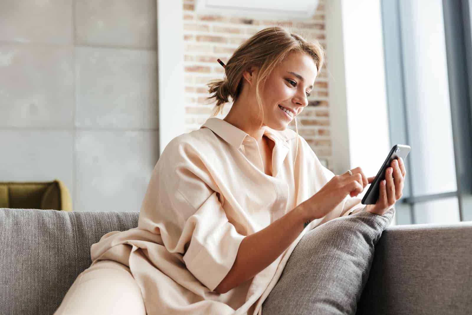 Une femme souriante aux cheveux attachés est assise sur le canapé et les touches du téléphone