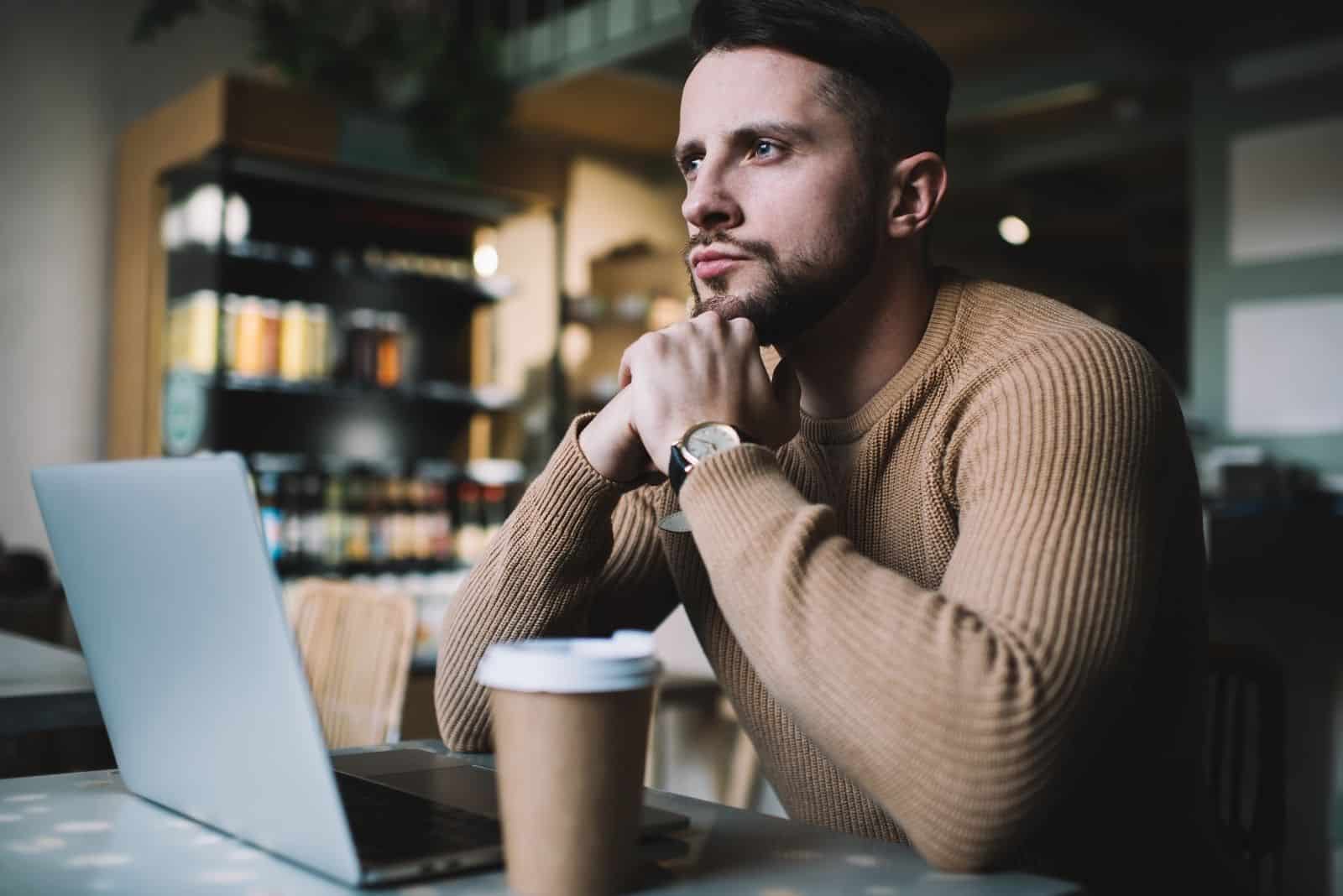 Homme barbu sérieux en tenue décontractée réfléchissant sur la solution de projet travaillant à distance dans l'intérieur du café avec une tasse à café jetable