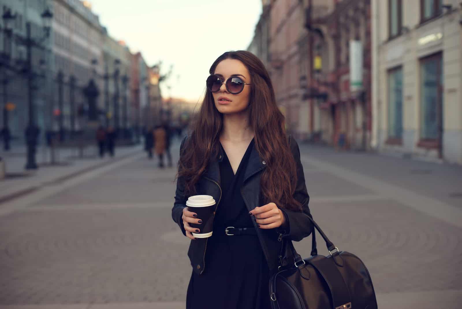 Une femme aux longs cheveux bruns avec des lunettes sur la tête se dresse dans la rue tenant un café à la main