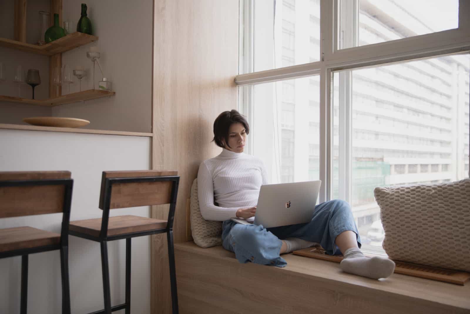 femme utilisant un ordinateur portable assis sur le rebord de la fenêtre