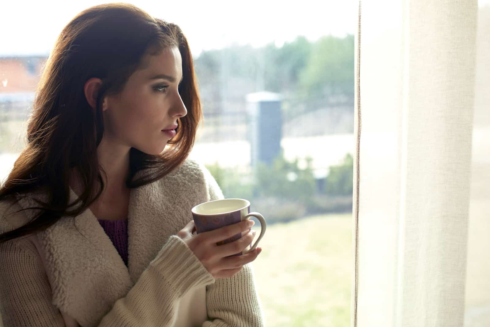 femme pensive tenant une tasse et debout près de la fenêtre
