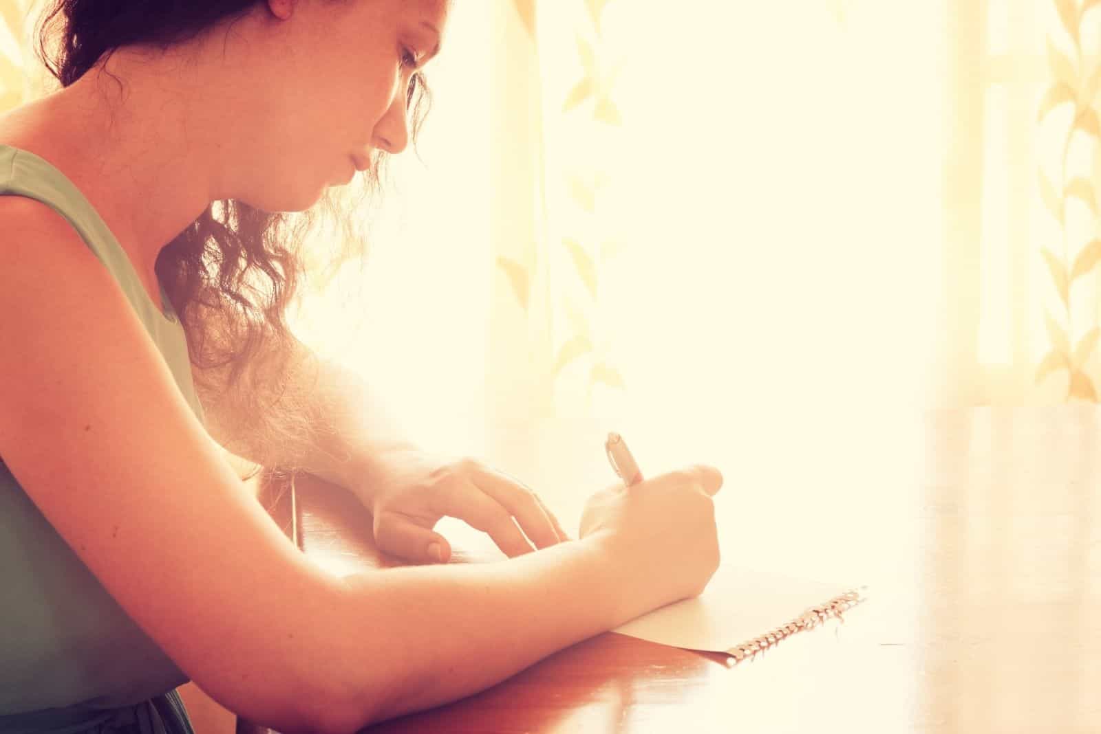 jeune femme assise près de la fenêtre et de l'écriture. image filtrée rétro. photographie avec la lumière naturelle des fenêtres