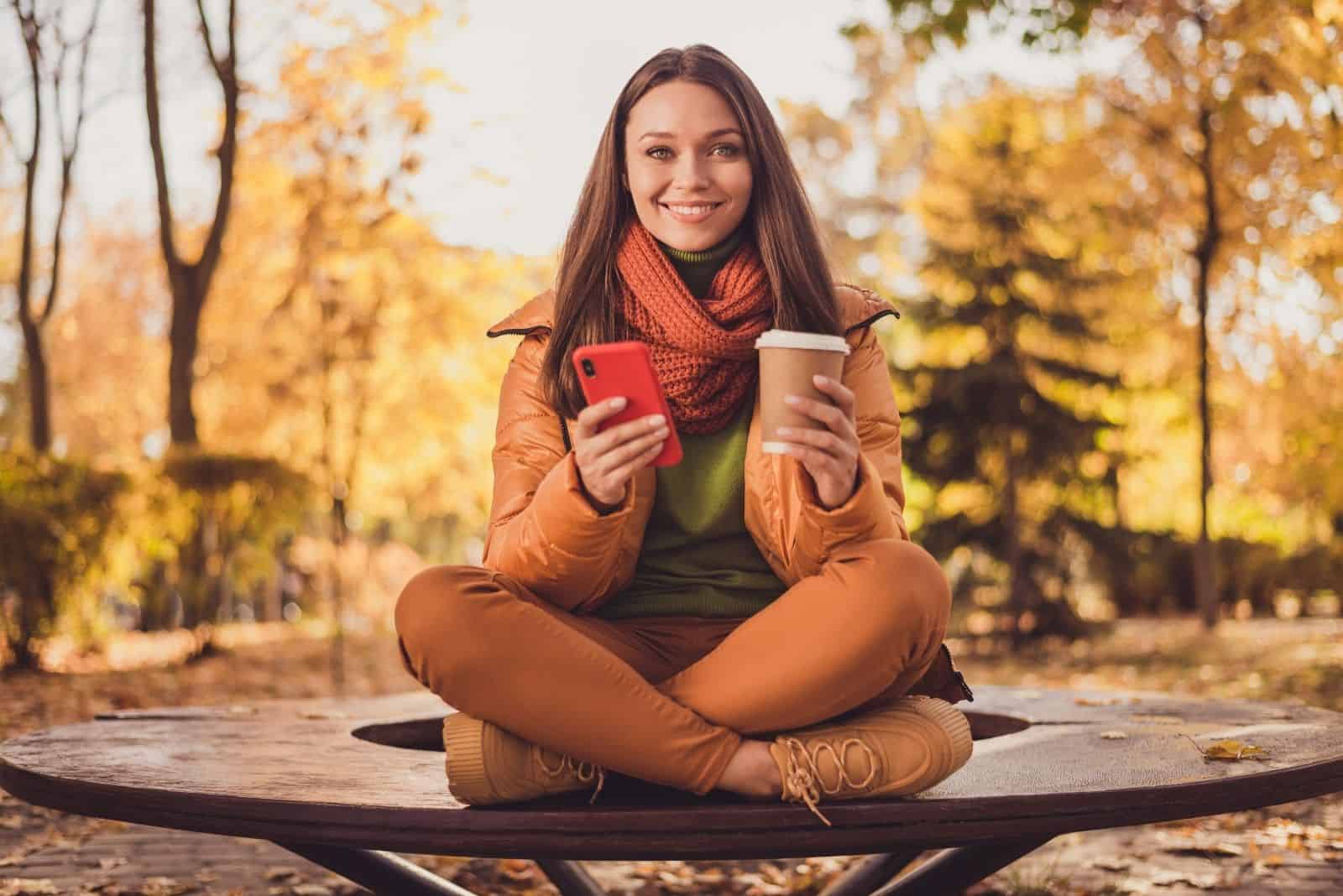 jolie dame souriante assise sur un banc tenant une tasse de téléphone après les cours à l'université bavardant avec un ami automne dans un parc écharpe verte col roulé orange pantalon coupe-vent baskets à l'extérieur