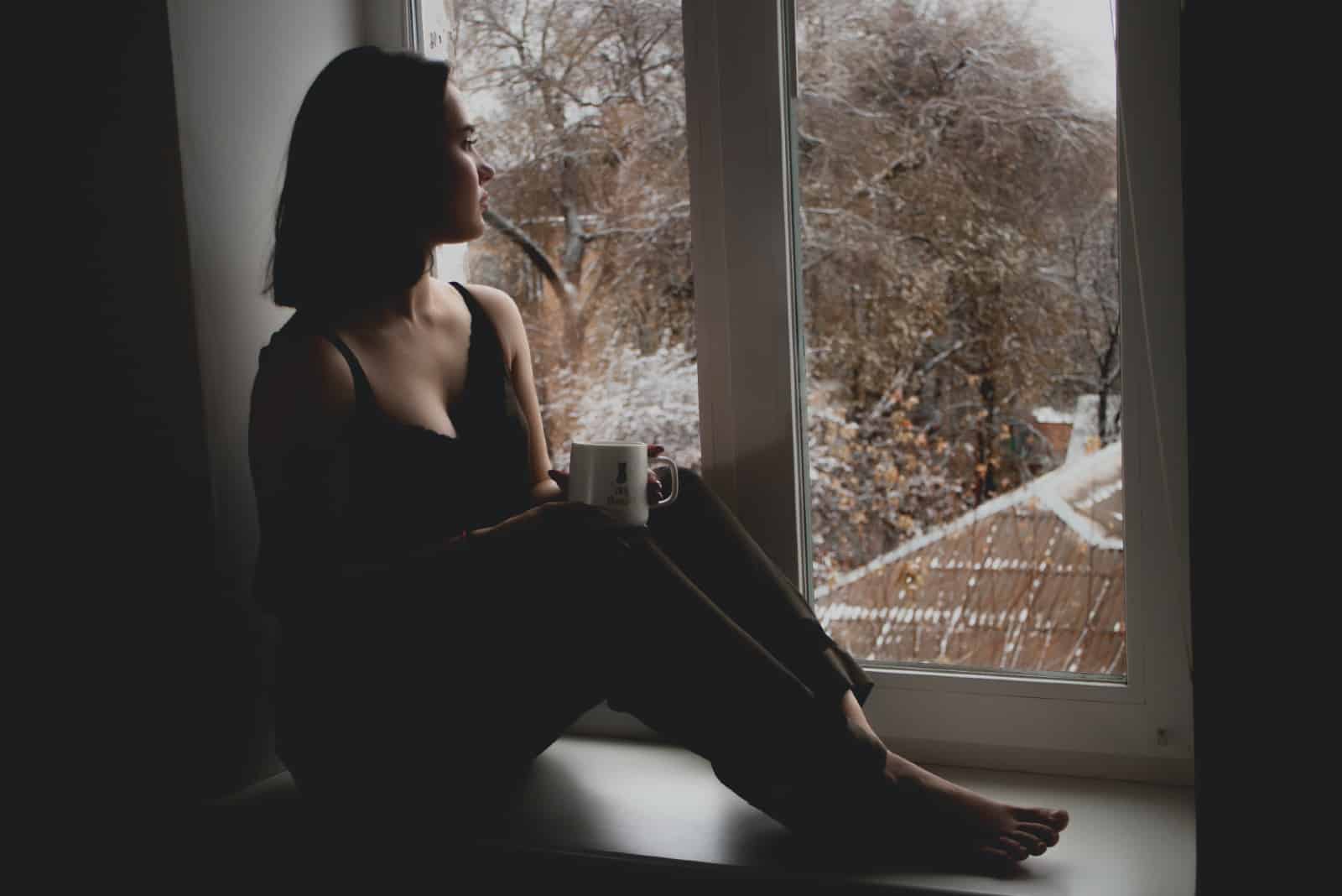 femme tenant une tasse assise sur le rebord d'une fenêtre