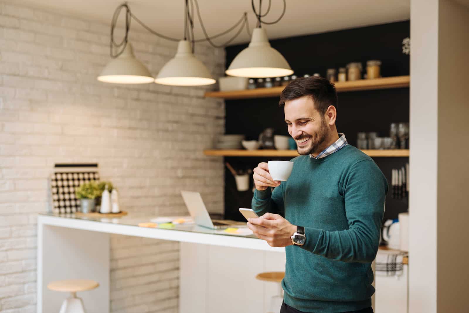 un homme souriant se tient dans la cuisine en train de boire du café et tenant un téléphone à la main