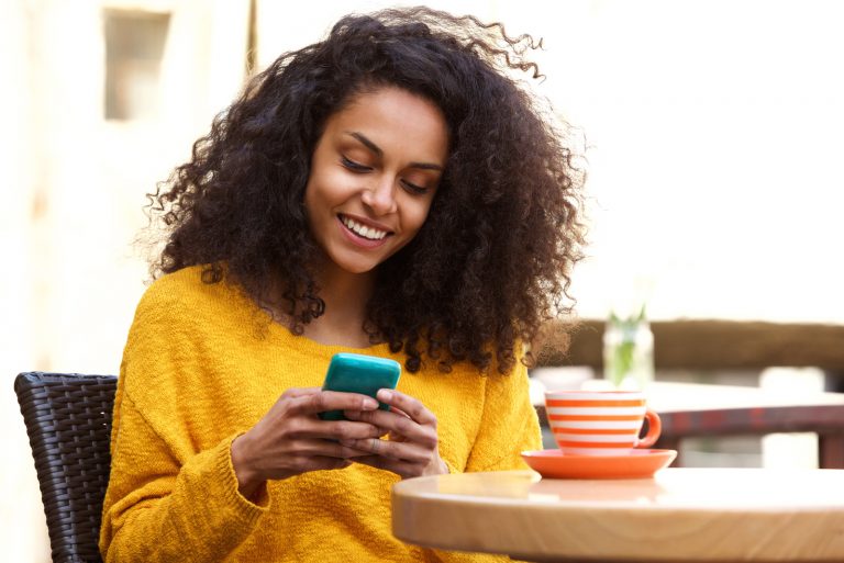 une femme souriante aux cheveux crépus assise à une table et tapant sur un téléphone