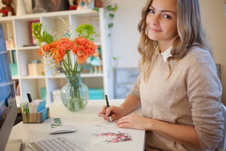 une femme aux cheveux blonds souriante est assise à une table et écrit